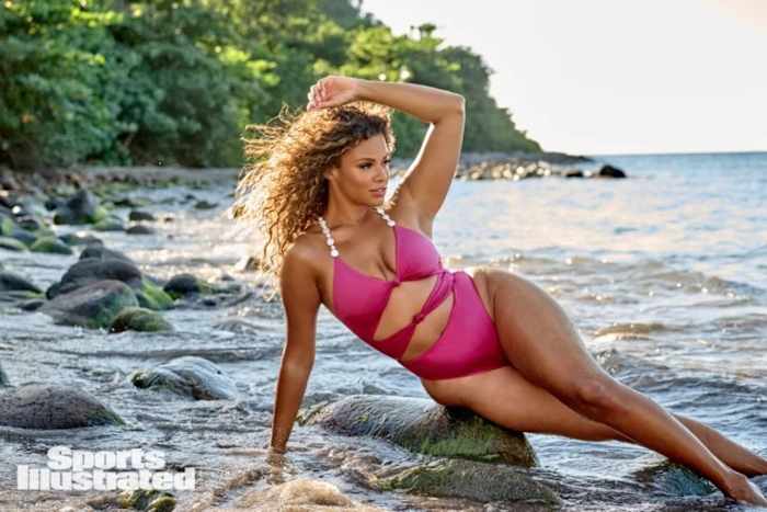 Kamie Crawford lounges on a rock in shallow water in a pink one-piece with cutouts.